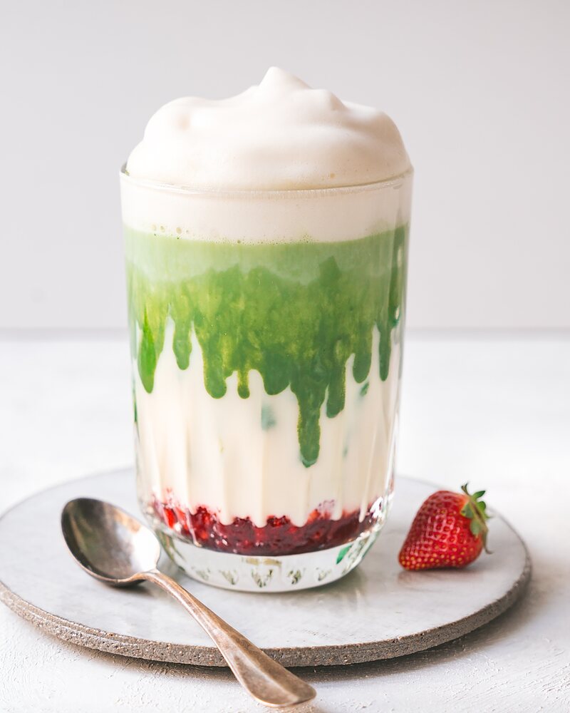 Matcha Latte mit Erdbeeren: Erdbeerpüree am Boden, darüber Milch, eine grüne Schicht Matcha und Milchschaum, daneben eine Erdbeere