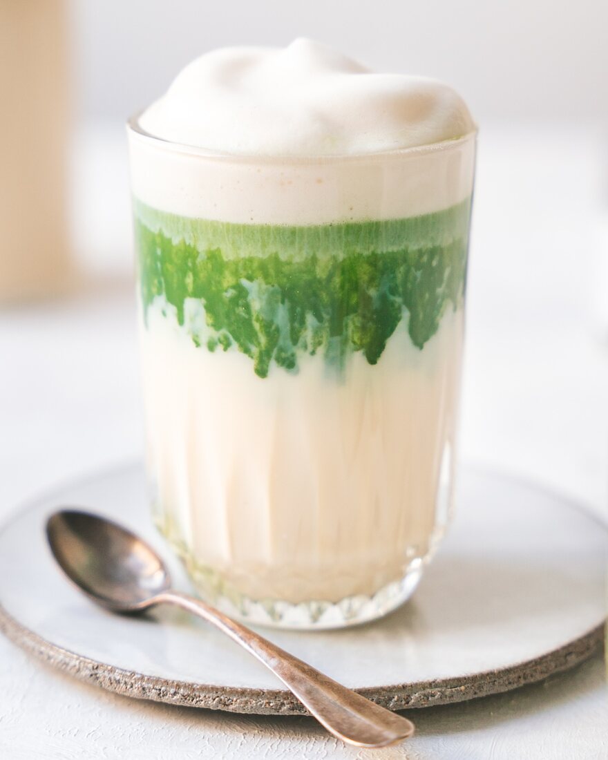 Matcha Latte im Glas: unten Milch, darüber eine leuchtend grüne Schicht Matcha und Milchschaum, daneben ein Löffel