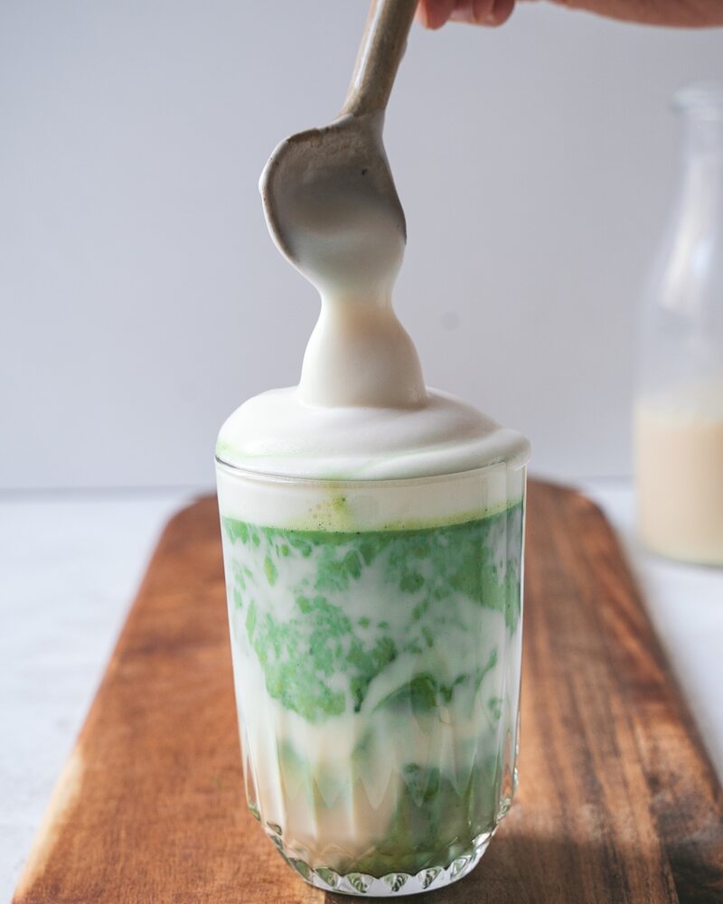 Milchschaum wird mit einem Löffel auf einen Matcha Latte im Glas gegeben