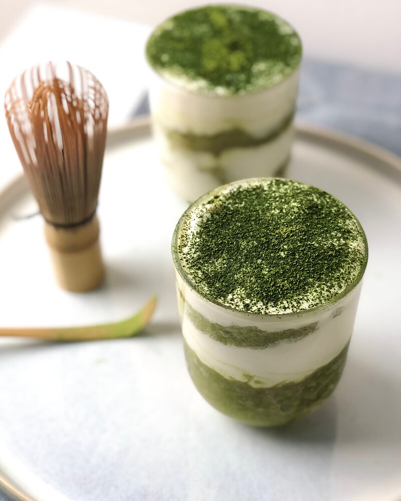 Matcha-Tiramisu in zwei Gläsern, mit Matcha-Pulver bestäubt, daneben ein Matchabesen und ein Bambuslöffel